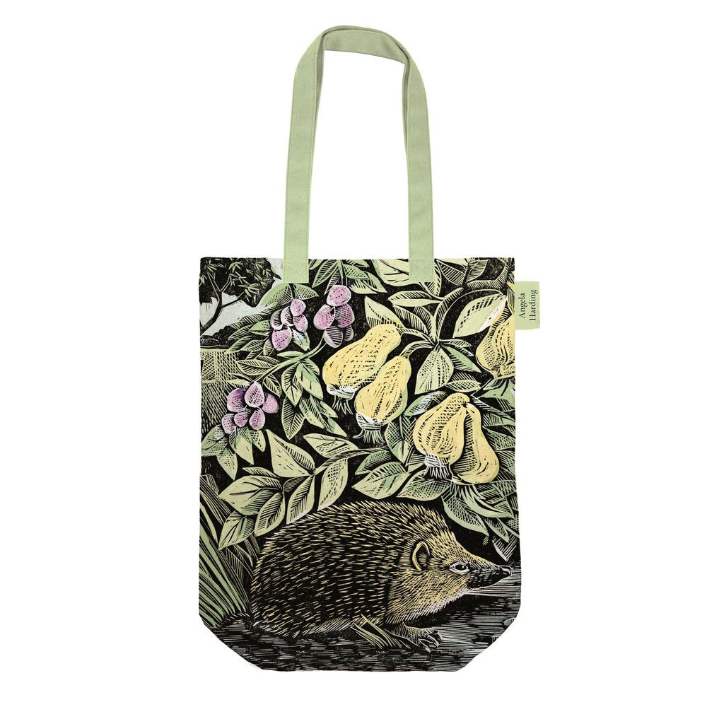 A canvas tote bag featuring an illustration of a hedgehog, pears, and purple flowers on a dark background, with light sage green handles and a small side tag.