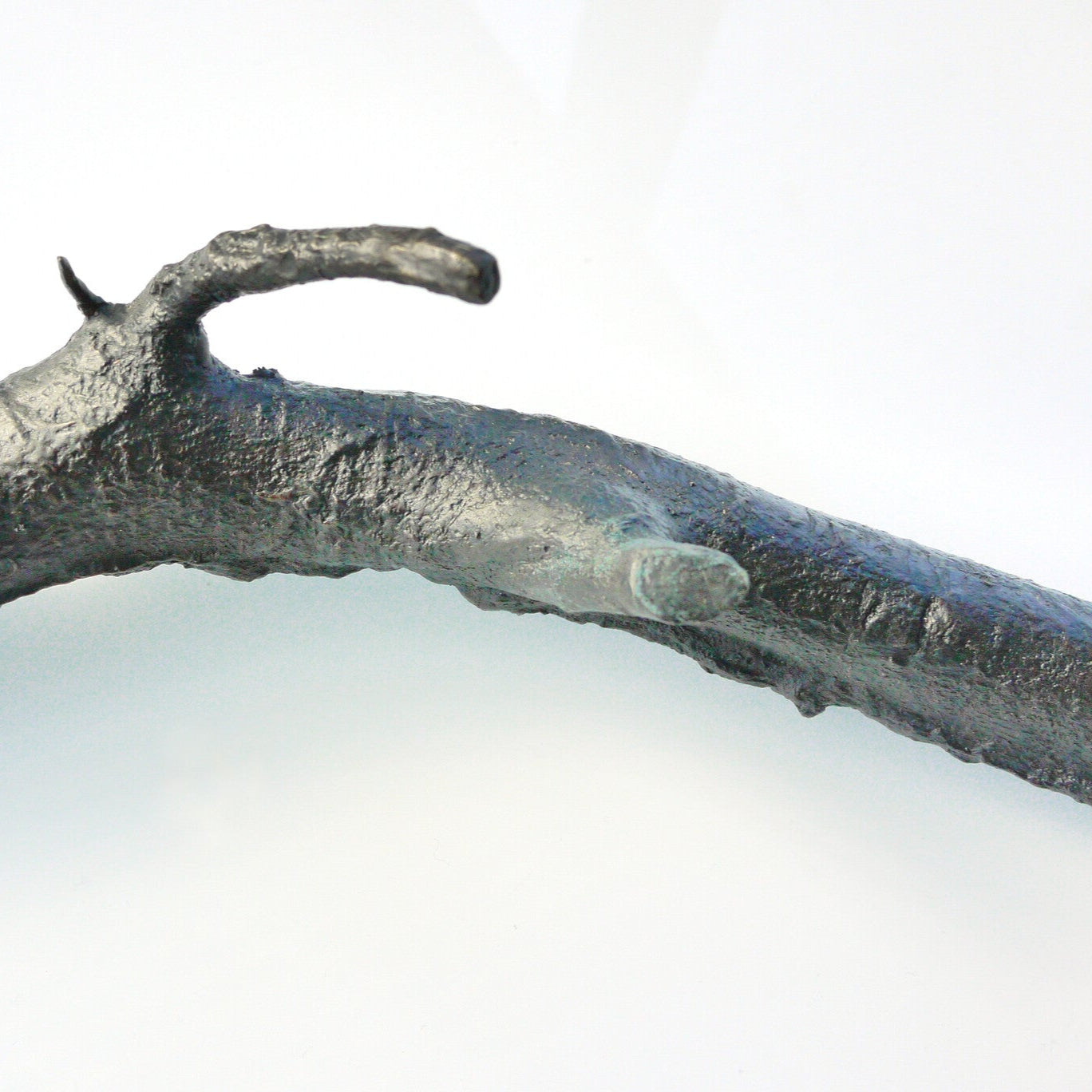 A textured, dark metallic branch-like sculpture lies on a light background. The piece features naturalistic details, including knots and curves, resembling a real twig, but is crafted from a solid material, giving it an artistic and modern appearance.