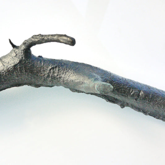 A textured, dark metallic branch-like sculpture lies on a light background. The piece features naturalistic details, including knots and curves, resembling a real twig, but is crafted from a solid material, giving it an artistic and modern appearance.