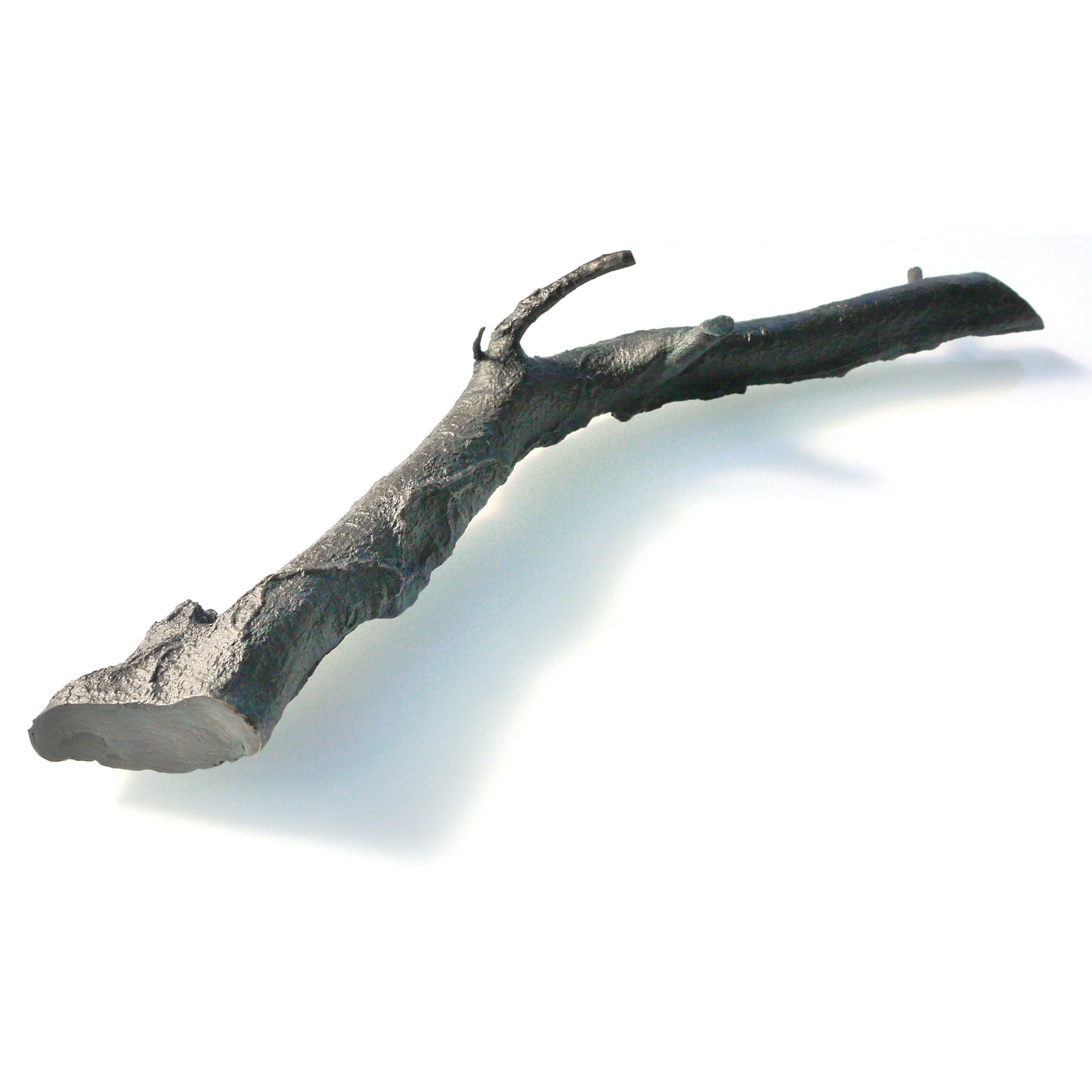 A textured, dark metallic branch-like sculpture lies on a light background. The piece features naturalistic details, including knots and curves, resembling a real twig, but is crafted from a solid material, giving it an artistic and modern appearance.