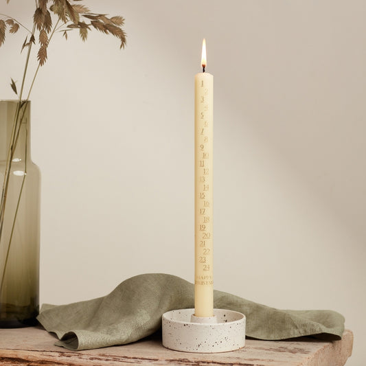 An ivory candle in a white ceramic holder, marked with numbers from 1 to 24, is lit and placed on a wooden surface next to a green vase and a red notebook.