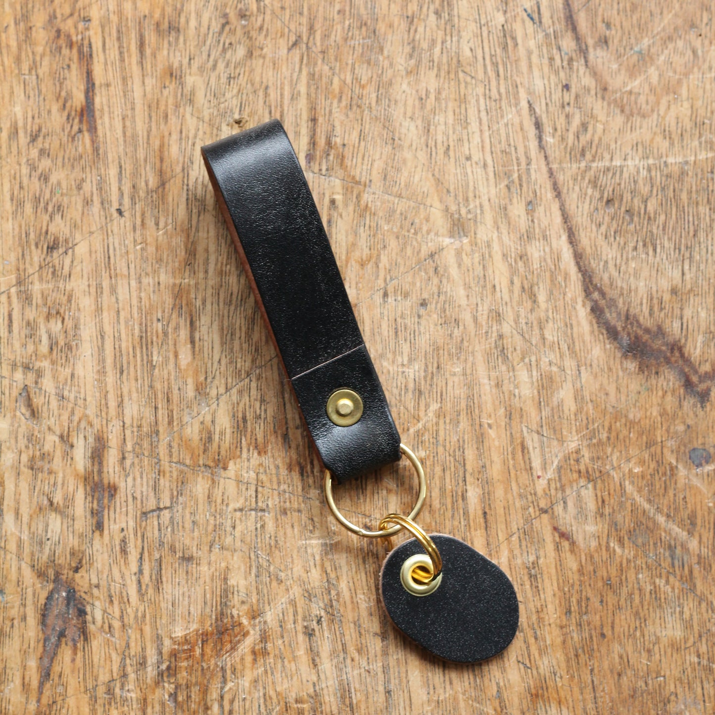 A black leather key fob with a brass ring and a brass tag, placed on a wooden surface.