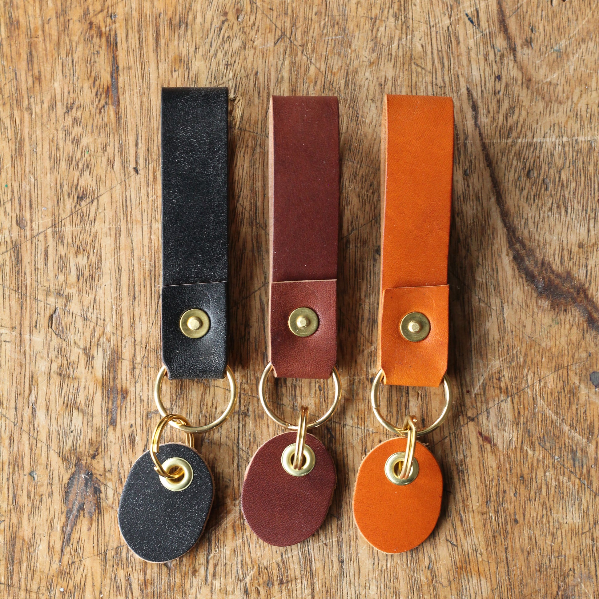 Three leather key fobs sitting next to each other on a wooden table, featuring brass rivets and a leather tag.