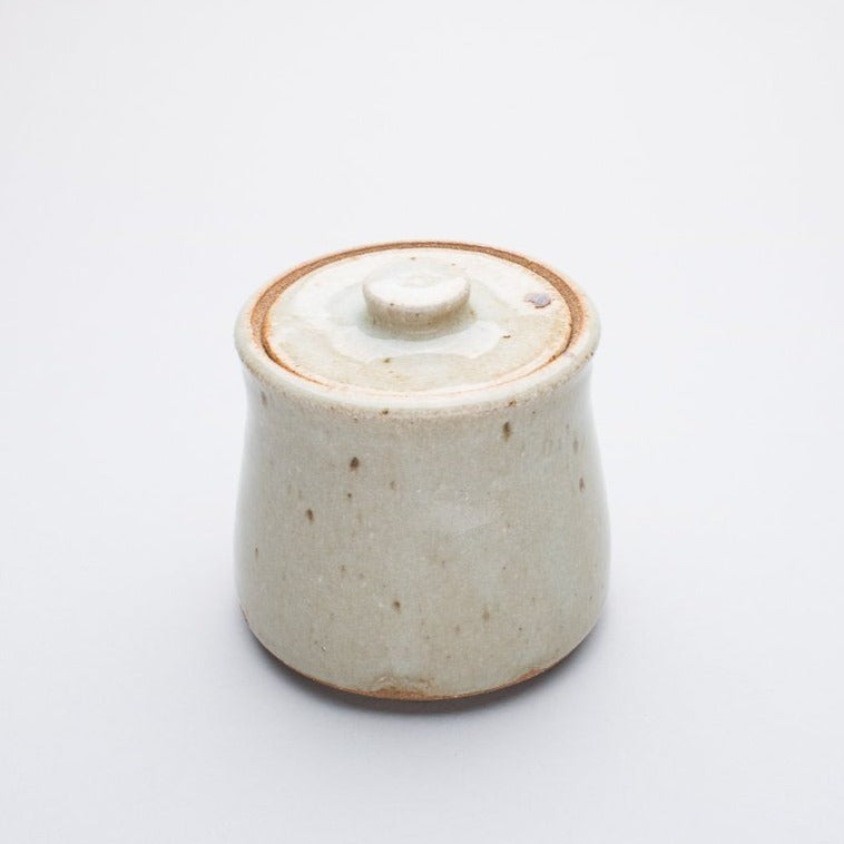 A beige ceramic honey jar with a lid, made of stoneware clay, with a creamy white glaze that may have a hint of blue.