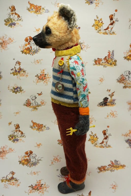 A handmade plush doll resembling a hyena stands against a floral background. It wears a striped jumper, patterned shirt, and red trousers. The doll has a fork accessory and button details, exuding a whimsical, vintage charm.