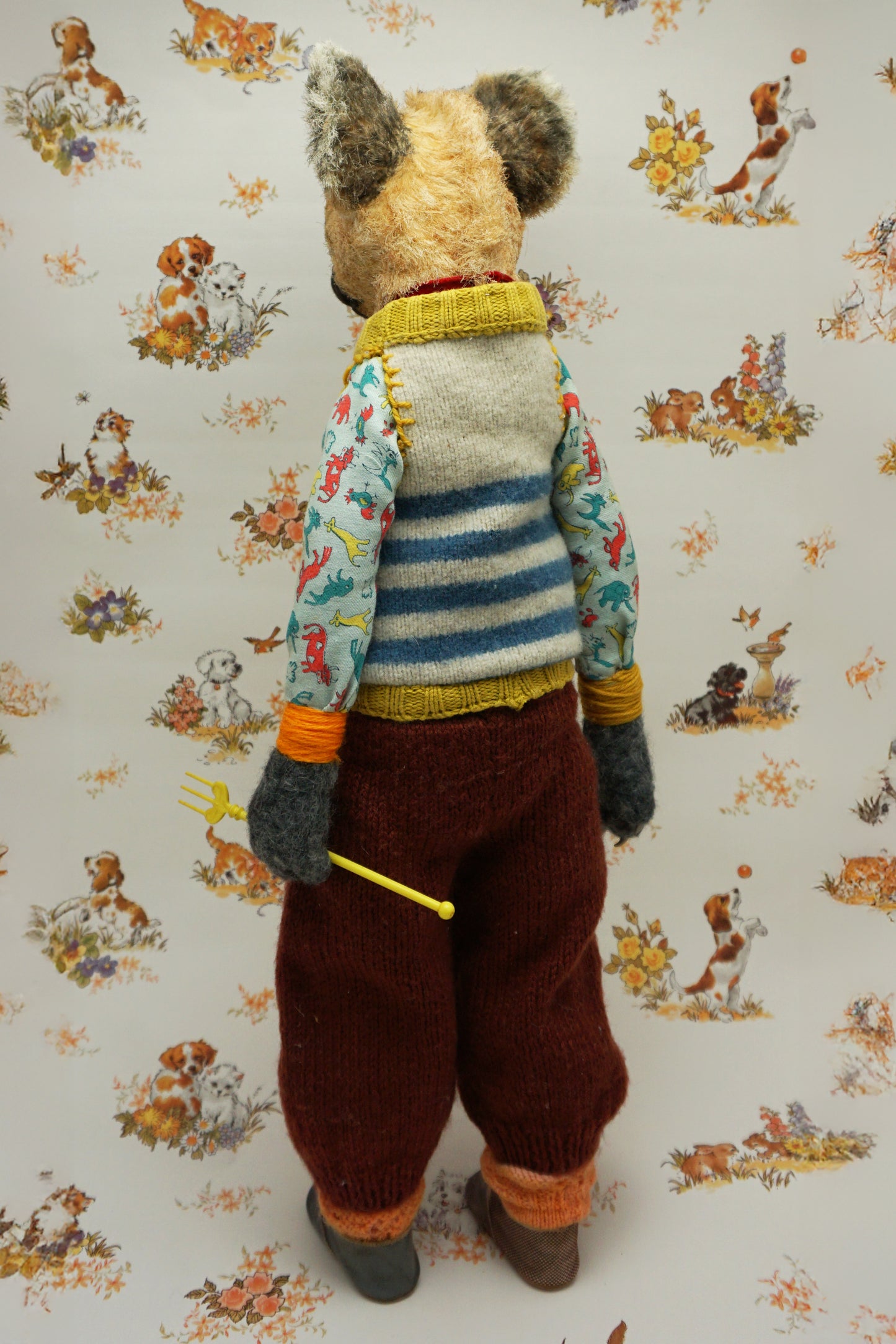 A handmade plush doll resembling a hyena stands against a floral background. It wears a striped jumper, patterned shirt, and red trousers. The doll has a fork accessory and button details, exuding a whimsical, vintage charm.