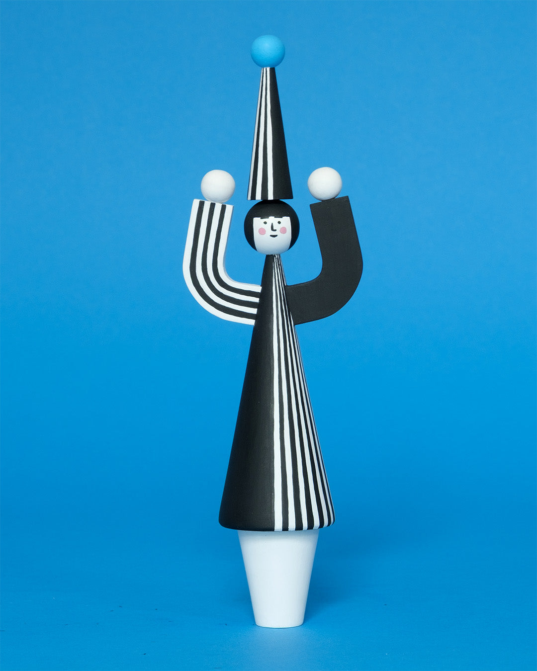 A wooden sculpture with a black and white stripey body with a matching headdress featuring a blue pompom on the top with white legs on a plain blue background.
