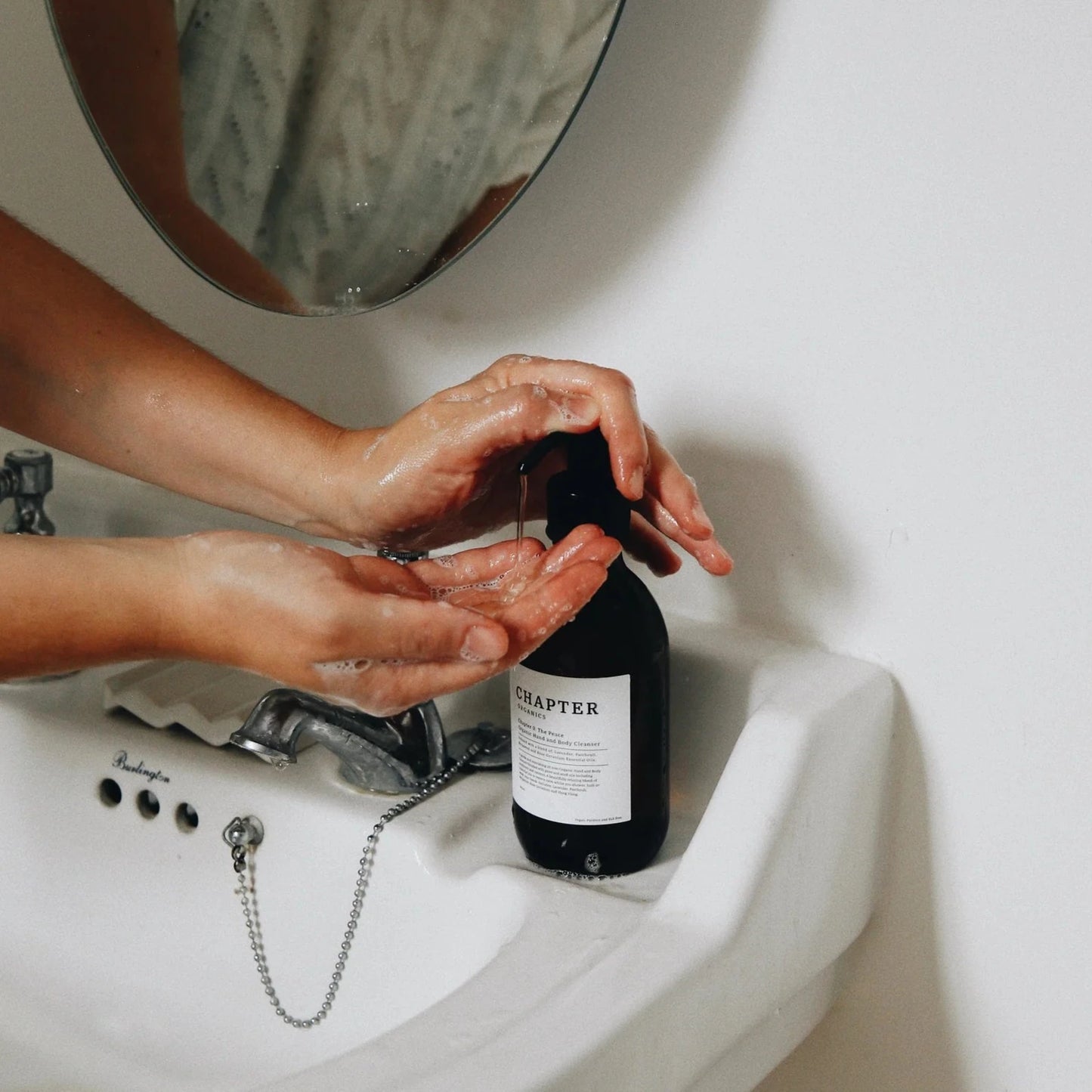 A person is washing their hands at a sink, using a dark bottle labelled "CHAPTER ORGANICS." The sink is white with a vintage-style tap, and a round mirror reflects the person's hands. The background is minimalistic and neutral.
