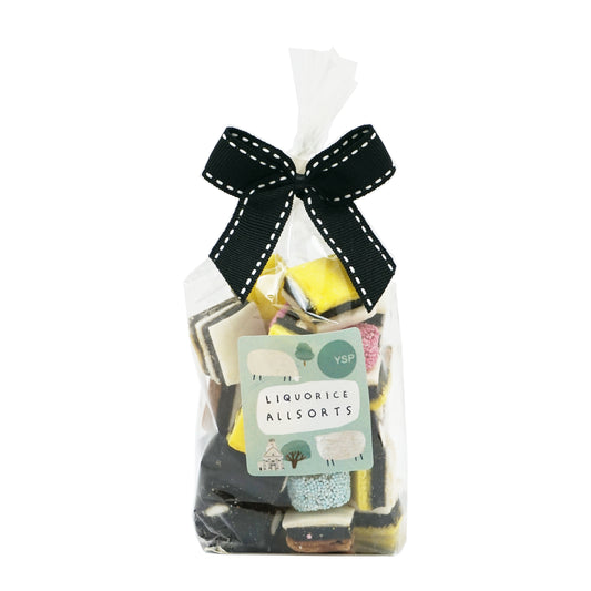 Packaged liquorice allsorts with a black ribbon on a white background. The label is green and features illustrations of sheep, trees and a building.