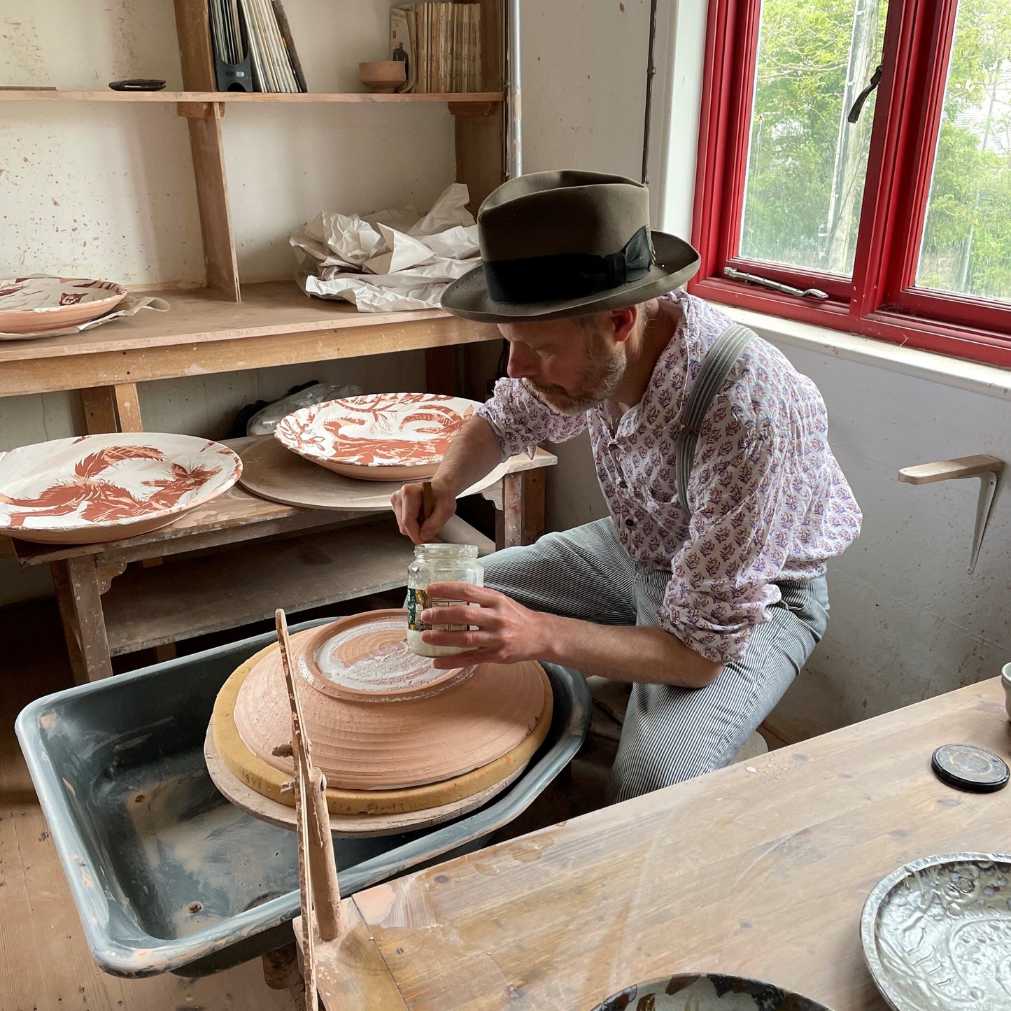 A white man sitting at a potters wheel wearing a hat and shirt waxing the bottom of a ceramic platter.