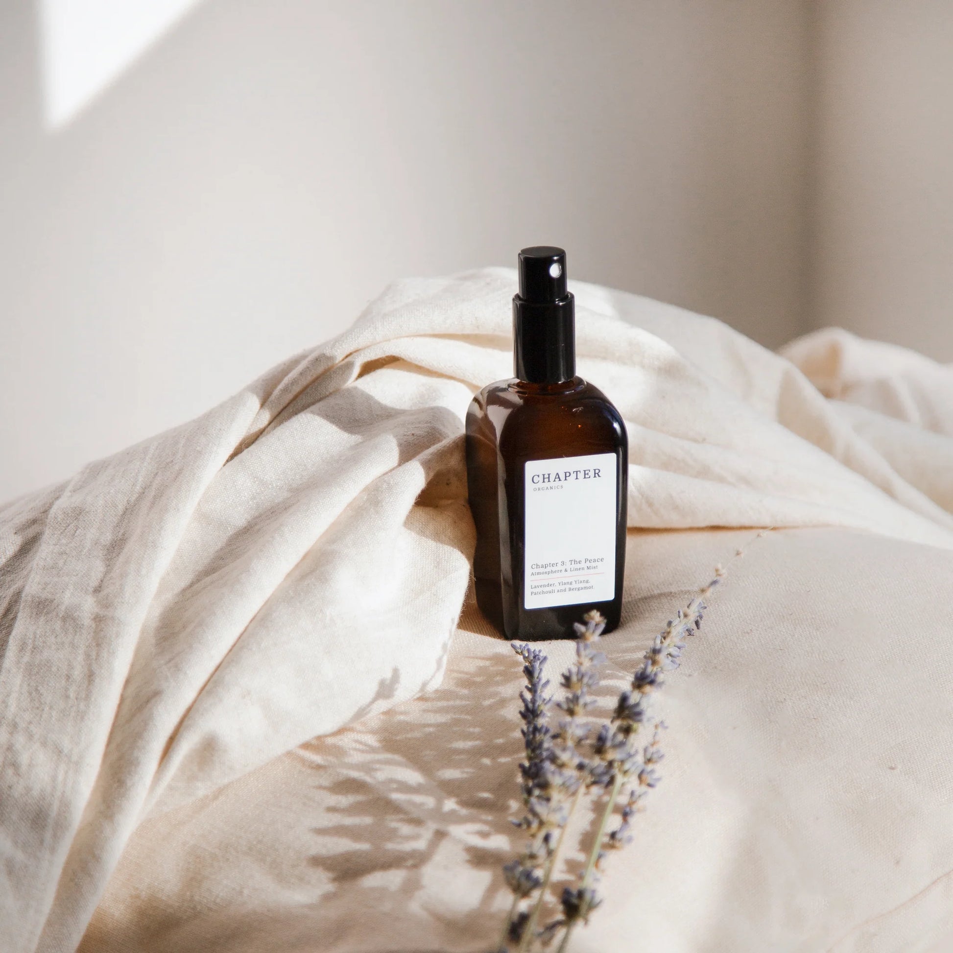 A brown glass spray bottle labelled "CHAPTER ORGANICS" sits on a soft, cream-coloured fabric. Nearby, a sprig of lavender adds a touch of colour, creating a serene and minimalist aesthetic in natural light.