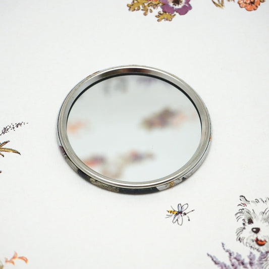 A round, silver-framed mirror lies on a white surface adorned with colourful floral patterns and a small dog illustration. The mirror reflects a blurred portion of the floral design.