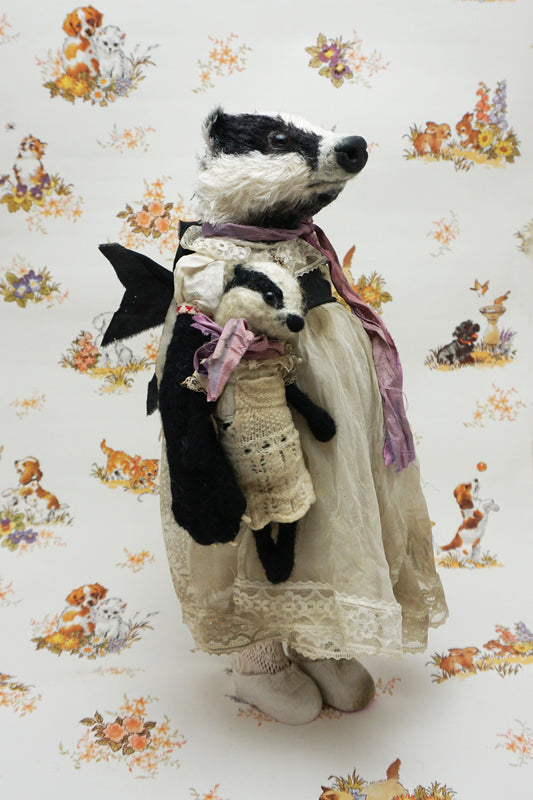 A plush badger toy dressed in a vintage-style lace dress and purple scarf stands holding a smaller badger toy in similar attire. The background features a whimsical pattern with animals and flowers.