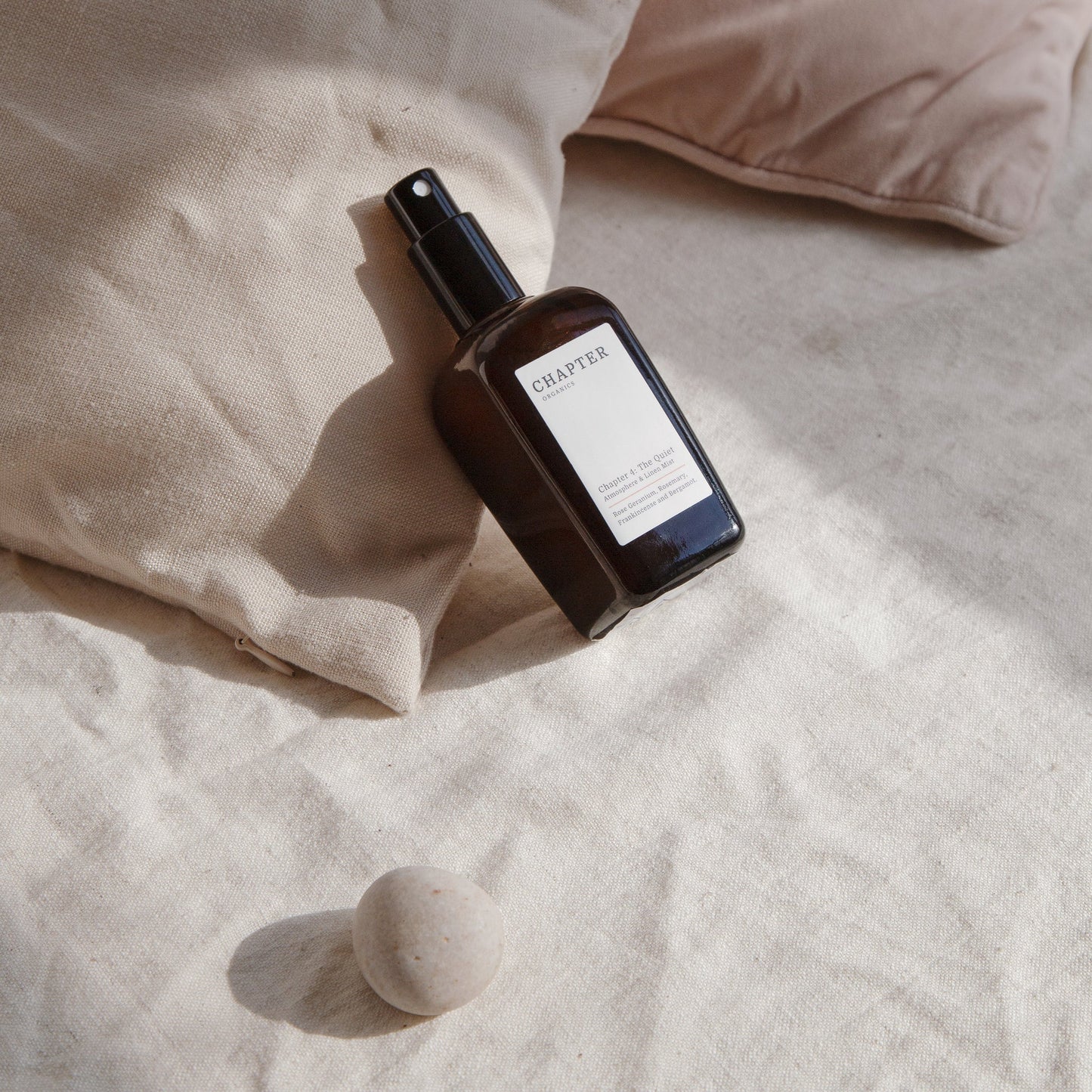 A brown glass spray bottle labelled "CHAPTER ORGANICS" rests on a light-coloured, textured fabric surface, accompanied by a smooth, round stone and two soft, neutral-toned cushions in the background. The scene conveys a serene, minimalist aesthetic.