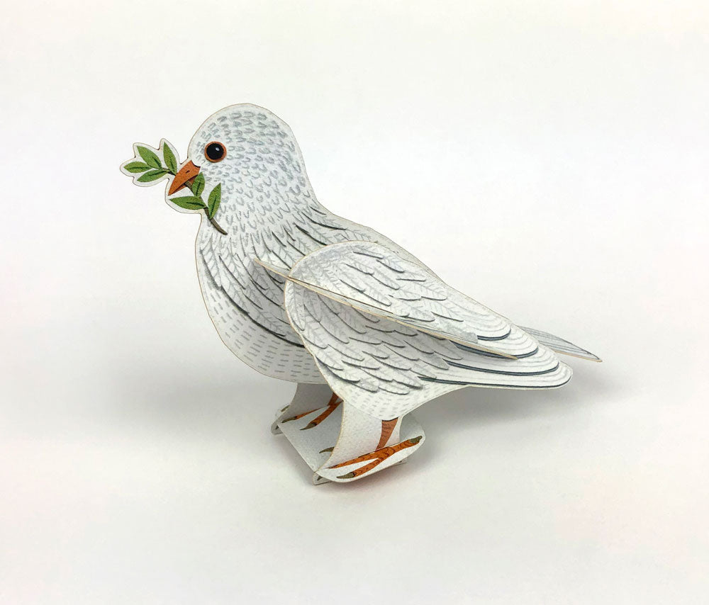 A three-dimensional pop-up card featuring a white dove holding an olive branch in its beak. The dove stands on a textured green nest, set against a soft blue background. The design is intricate, showcasing detailed feather patterns.