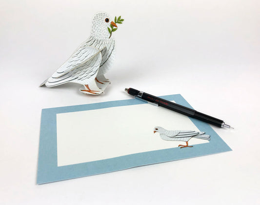 A three-dimensional pop-up card featuring a white dove holding an olive branch in its beak. The dove stands on a textured green nest, set against a soft blue background. The design is intricate, showcasing detailed feather patterns.