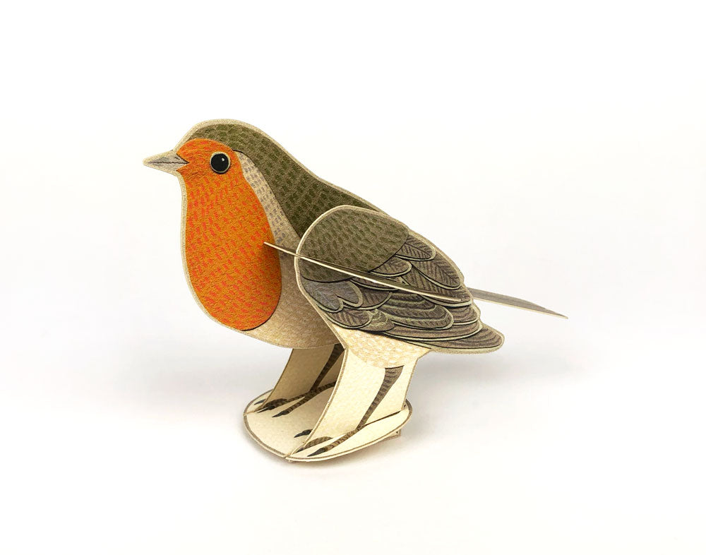 A three-dimensional paper model of a robin with an orange breast and detailed feather patterns, standing on a green card featuring holly leaves and berries. The background is plain white, highlighting the vibrant colours of the bird and card.