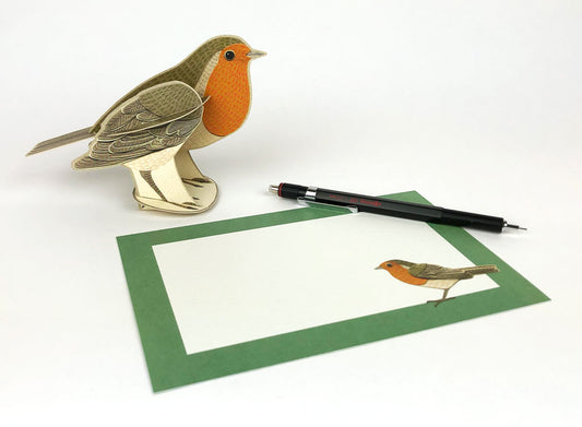 A three-dimensional paper model of a robin with an orange breast and detailed feather patterns, standing on a green card featuring holly leaves and berries. The background is plain white, highlighting the vibrant colours of the bird and card.