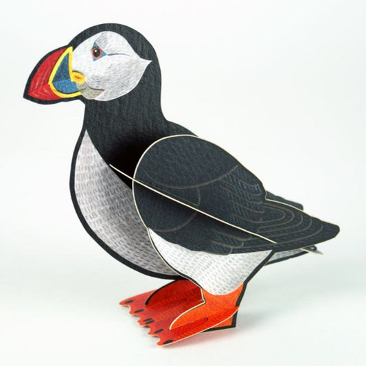 A colourful paper model of a puffin, featuring a black and white body, bright orange feet, and a vibrant beak. The model is positioned upright against a plain white background, showcasing its detailed illustration and playful design.
