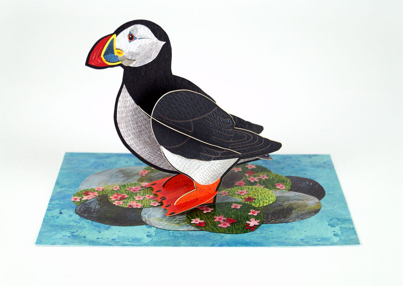 A colourful paper model of a puffin stands upright on a textured blue background, surrounded by green rocks and pink flowers. The puffin features a black and white body, bright orange feet, and a distinctive multicoloured beak.