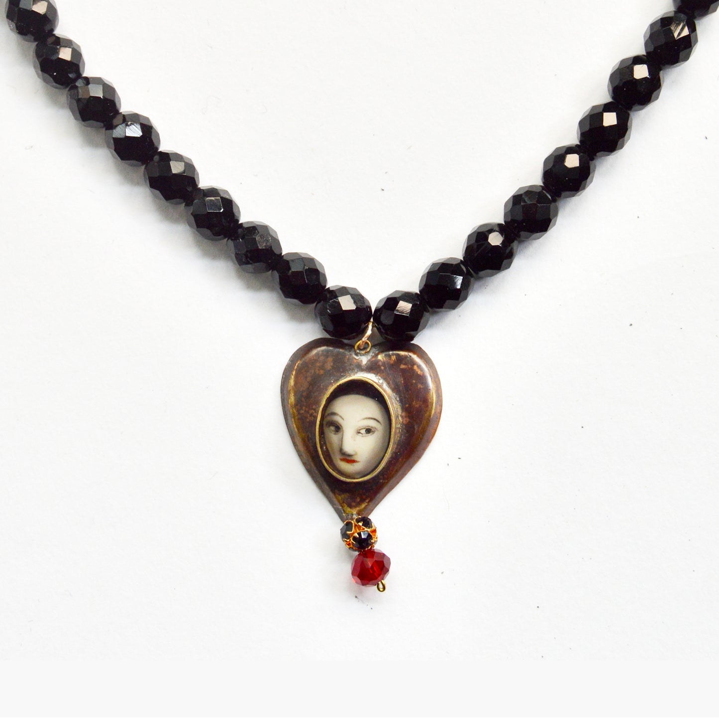 A close up detail showing the heart pendant on the beaded chain. There is a pale women's face inside the middle and two bead dangling underneath, one red one black.