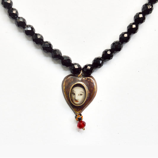 A close up detail showing the heart pendant on the beaded chain. There is a pale women's face inside the middle and two bead dangling underneath, one red one black.