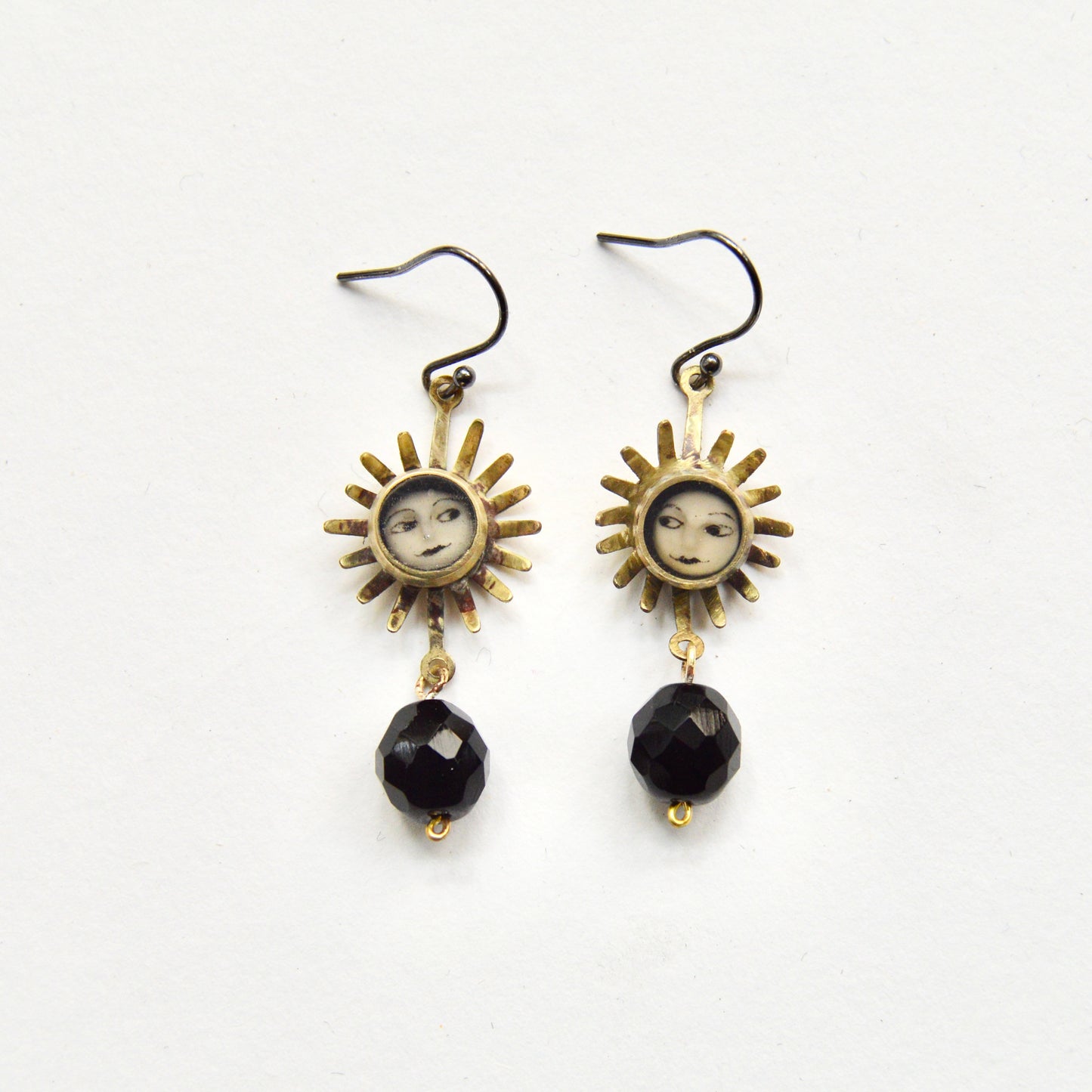 A pair of hanging earrings featuring a sun with a painted face inset on each earring. Under the sun dangles a single black bead.