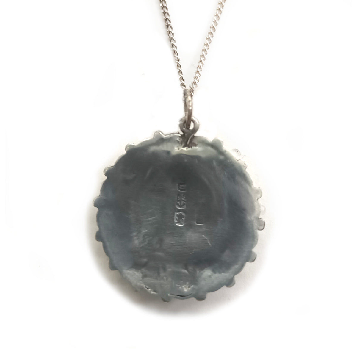The back of a pendant, showing a shiny smooth silver surface attached to a silver fine chain. The back appears to feature some hallmarking symbols.