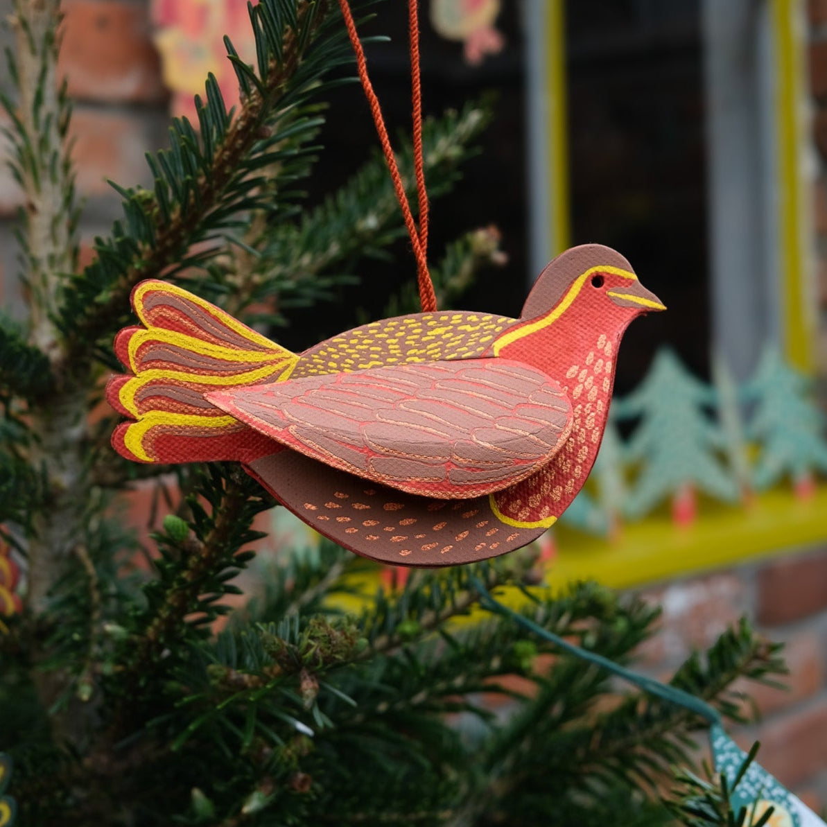 Decorative bird ornament on a Christmas tree with a window and brick wall in the background.