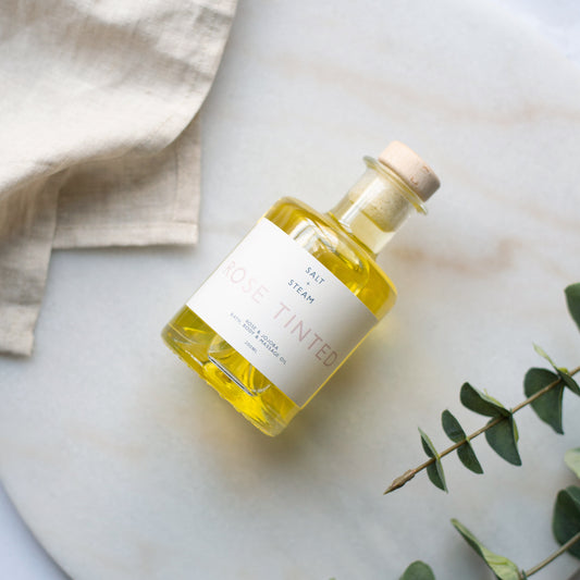 Bottle of 'Salt & Steam' rose tinted oil on a marble surface with a beige towel and green leaves.