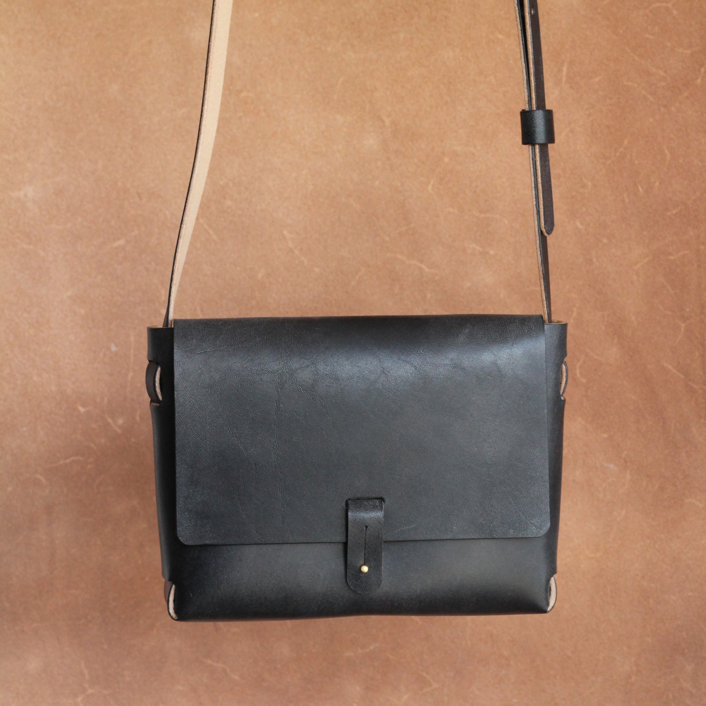 A black leather handbag with a long shoulder strap hanging against a brown background. The bag is simple is design and elegant, and features a brass stud closure.