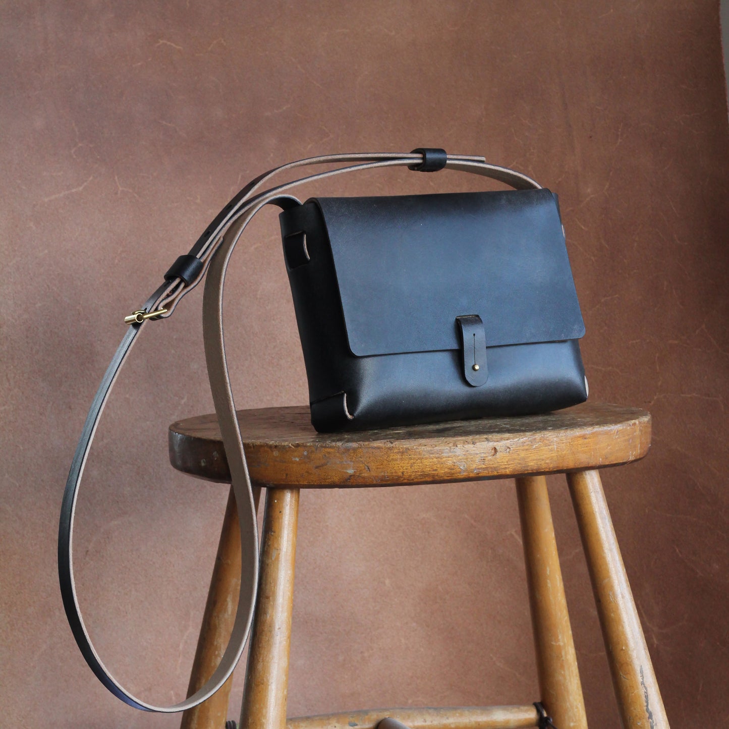 A black leather handbag with a long shoulder strap placed on a wooden stool, against a brown background. The bag is simple is design and elegant.
