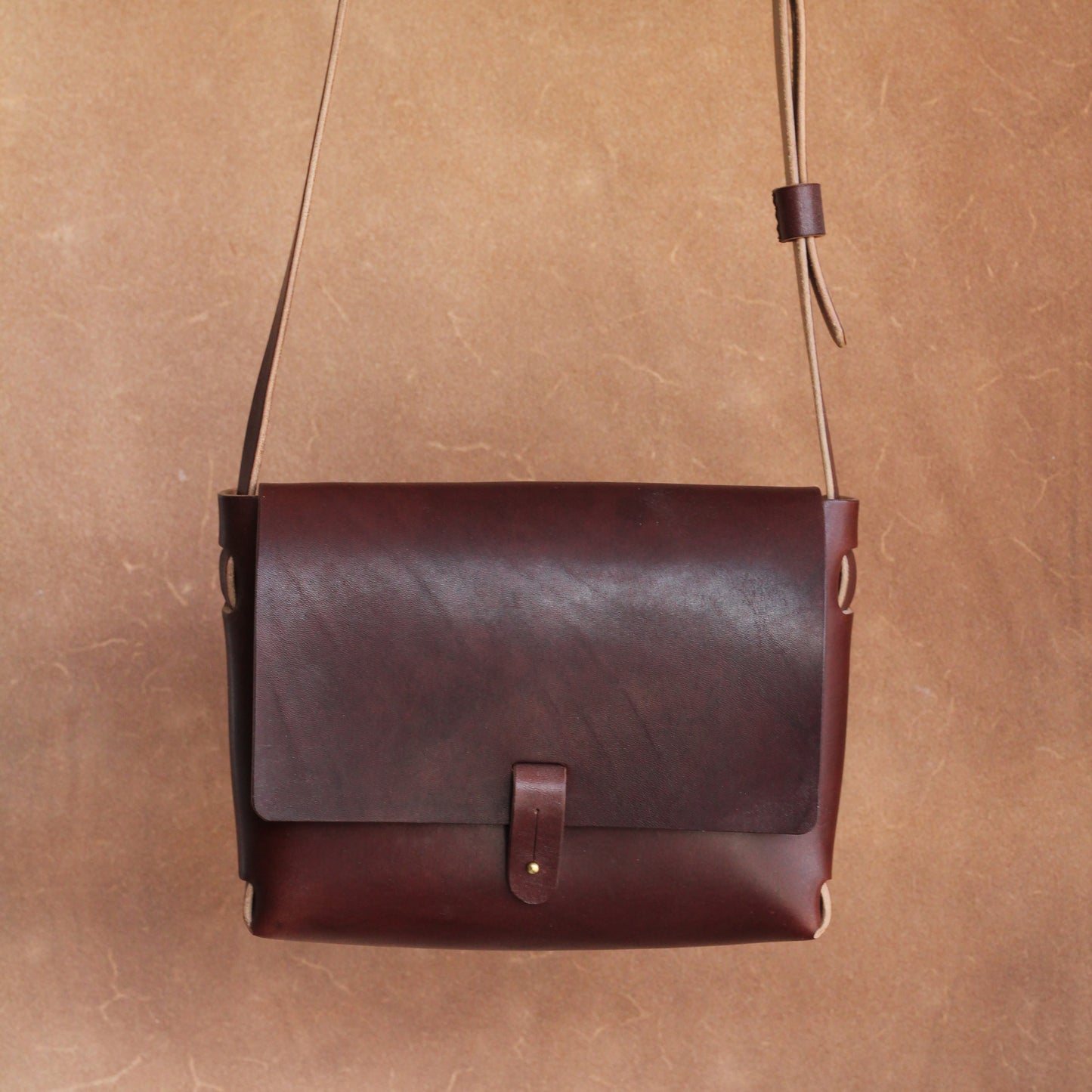 A brown leather handbag with a long shoulder strap hanging against a brown background. The bag is simple is design and elegant, and features a brass stud closure.