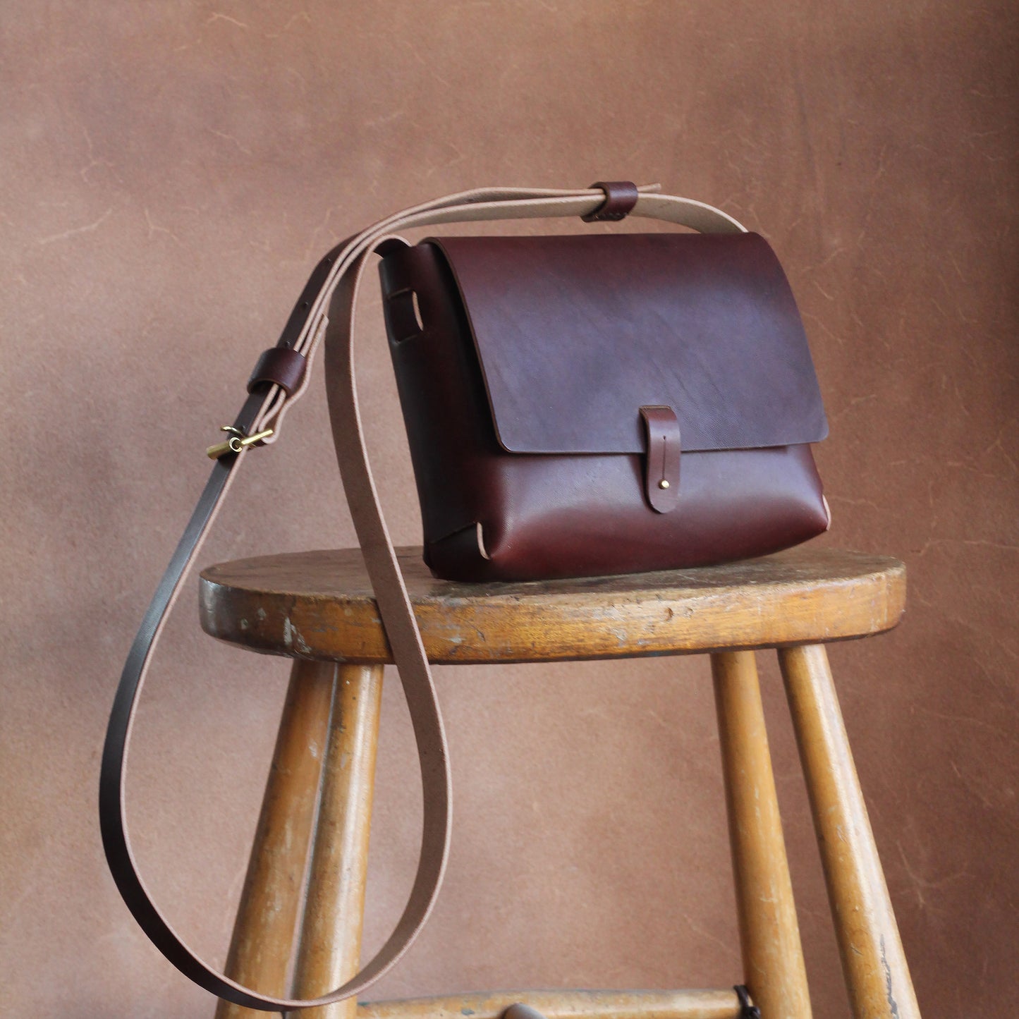 A brown leather handbag with a long shoulder strap placed on a wooden stool, against a brown background. The bag is simple is design and elegant.
