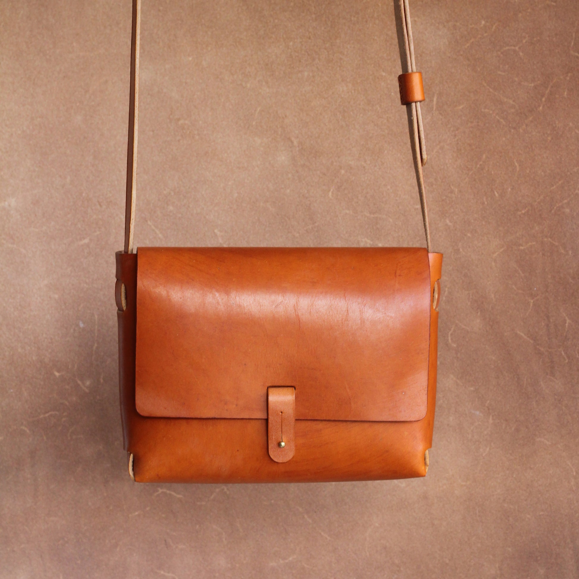 A tan leather handbag with a long shoulder strap hanging against a brown background. The bag is simple is design and elegant, and features a brass stud closure.