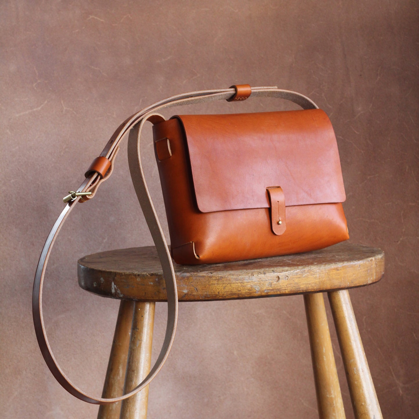 A tan leather handbag with a long shoulder strap placed on a wooden stool, against a brown background. The bag is simple is design and elegant.