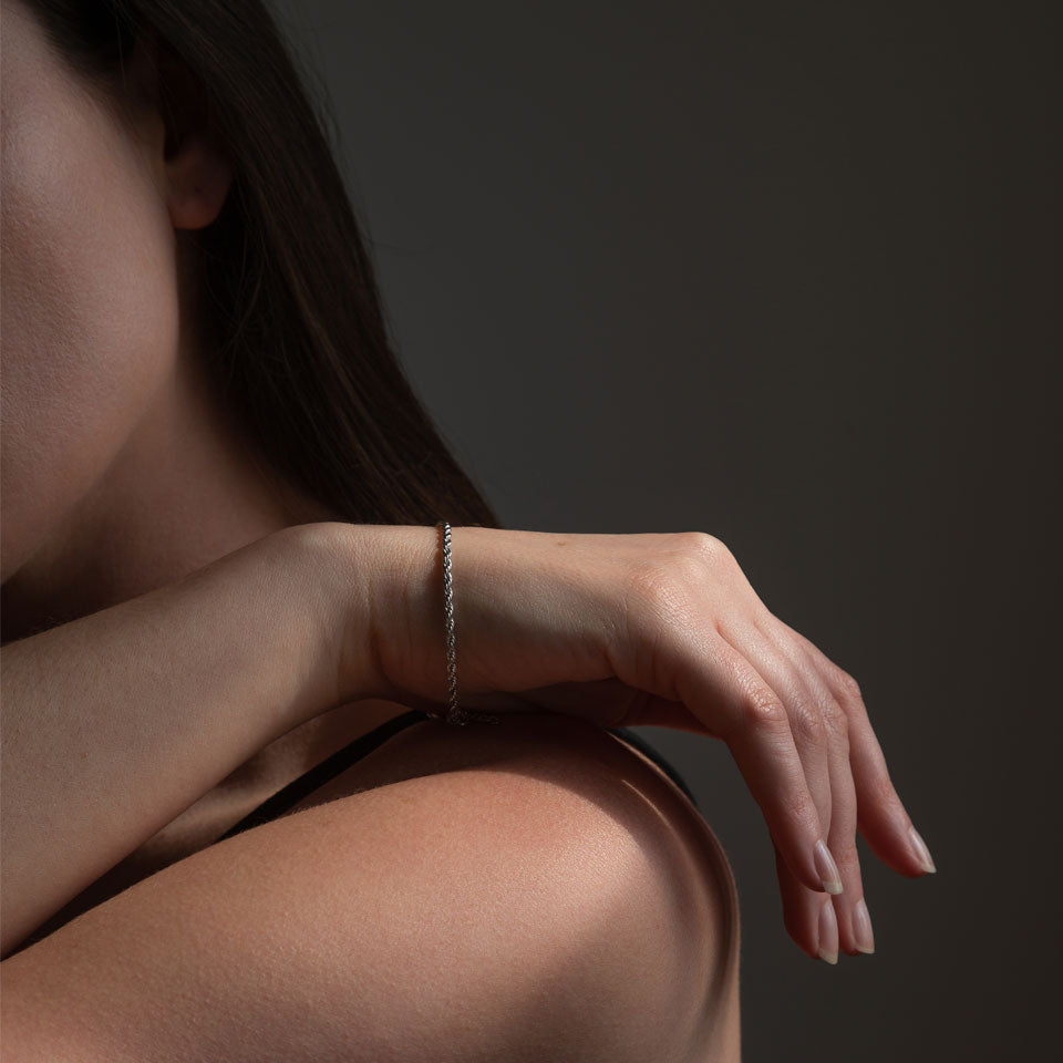 A person wearing a silver rope bracelet on their wrist, with the bracelet in focus and the rest of the image relatively dark.