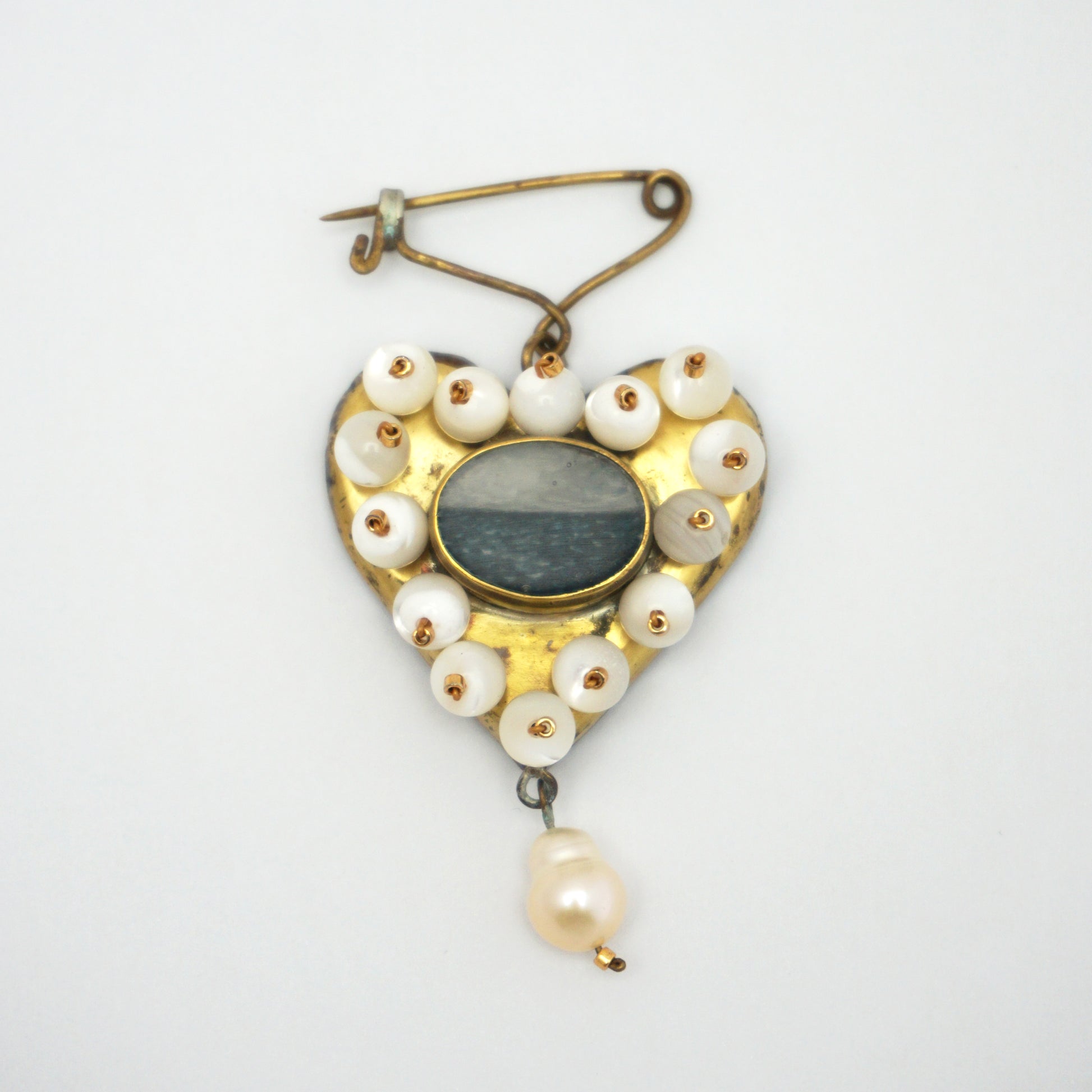 Gold coloured heart-shaped brooch with pearls on the outer edge and a central hand painted ocean insert. The brooch is on a plain white background and features a single pearl dangling from the bottom of the heart.