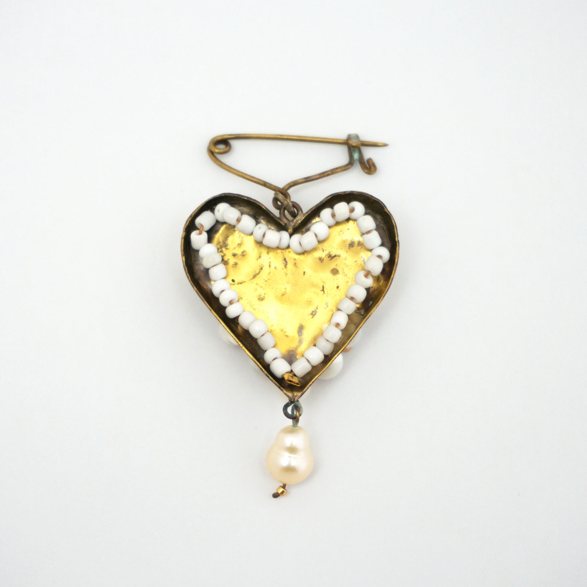 The back of a gold coloured heart-shaped brooch with pearls on the outer edge. The material is smooth and shiny.