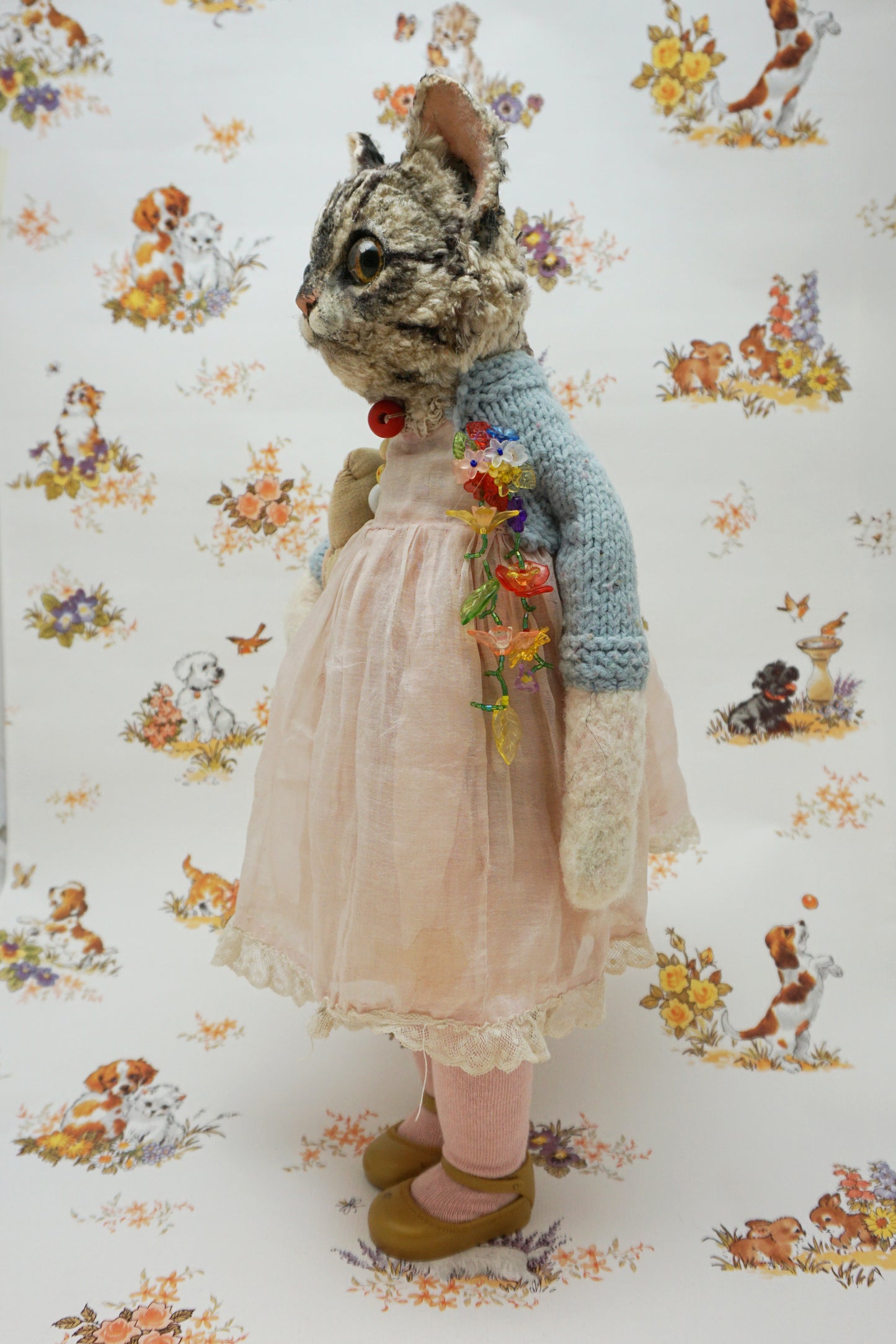 A plush grey tabby cat stands upright, wearing a pink dress, blue cardigan, and holding a fish soft toy. It has large eyes and a red bead for a collar. The background features a floral and animal pattern with small dogs and flowers.