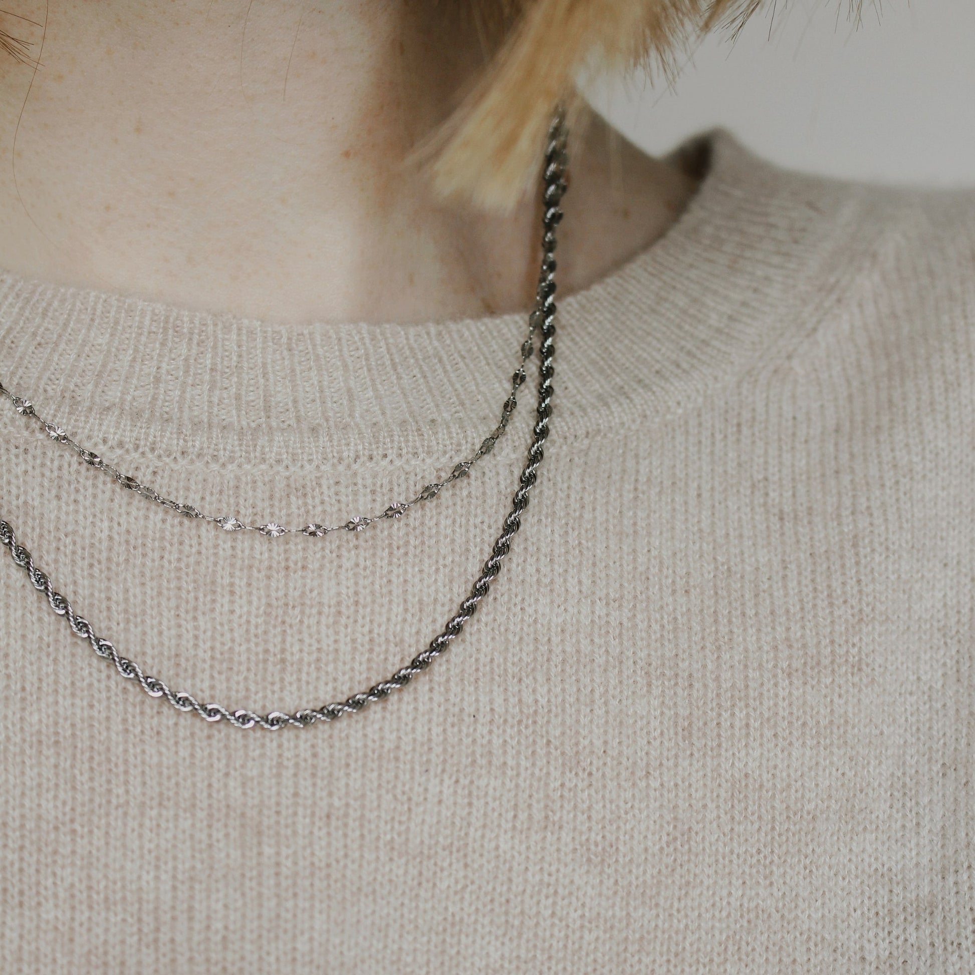 One necklace that gives the appearance of two silver necklaces layered on a person wearing a beige jumper.