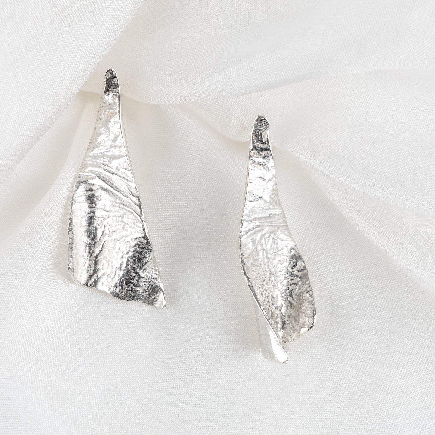 A pair of triangular silver textured earrings laid on a white fabric surface.