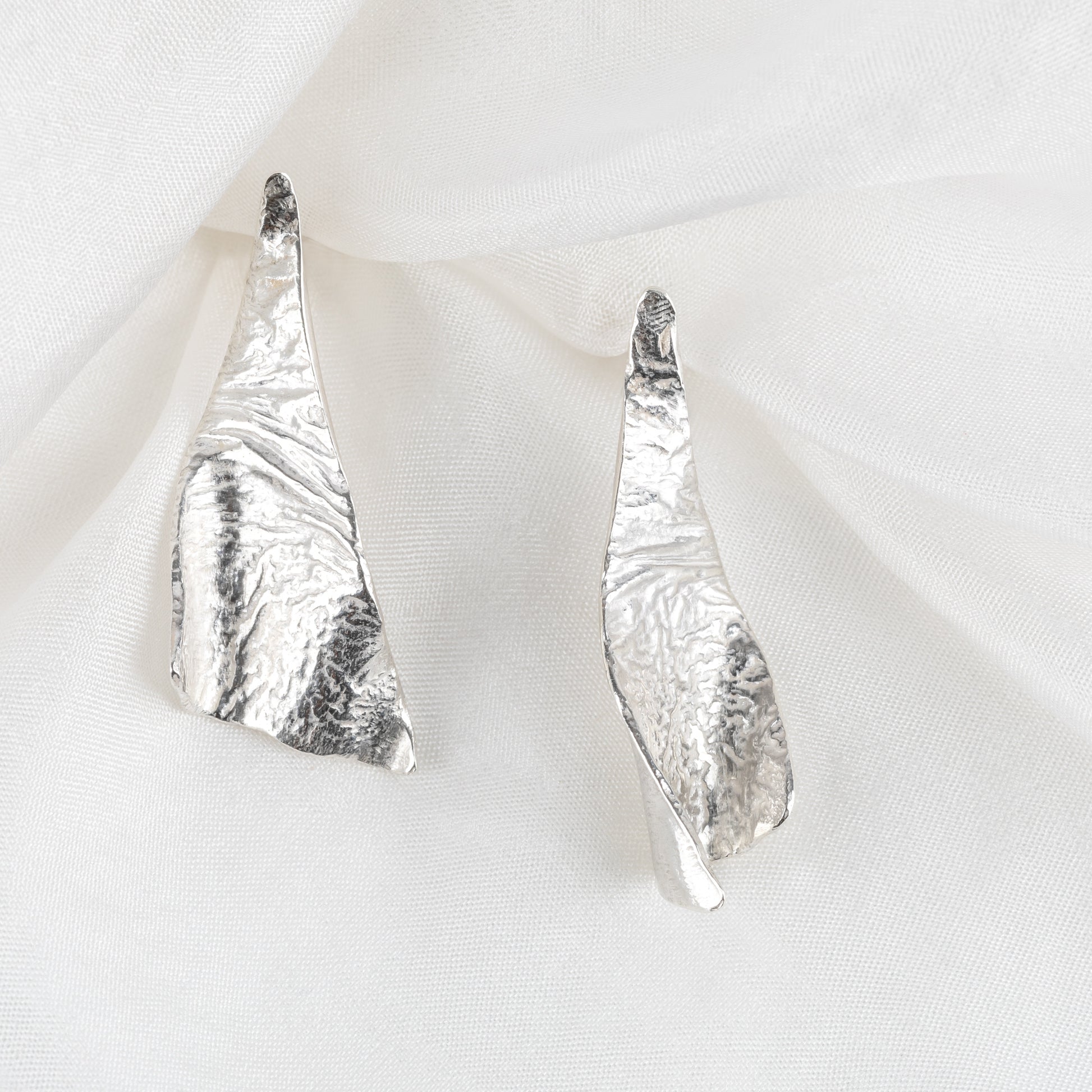 A pair of triangular silver textured earrings laid on a white fabric surface.