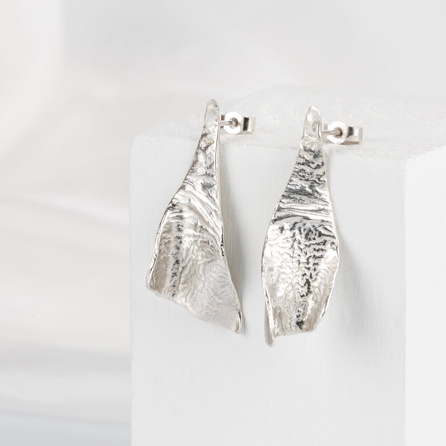 A pair of triangular silver textured earrings laid on a white fabric surface.