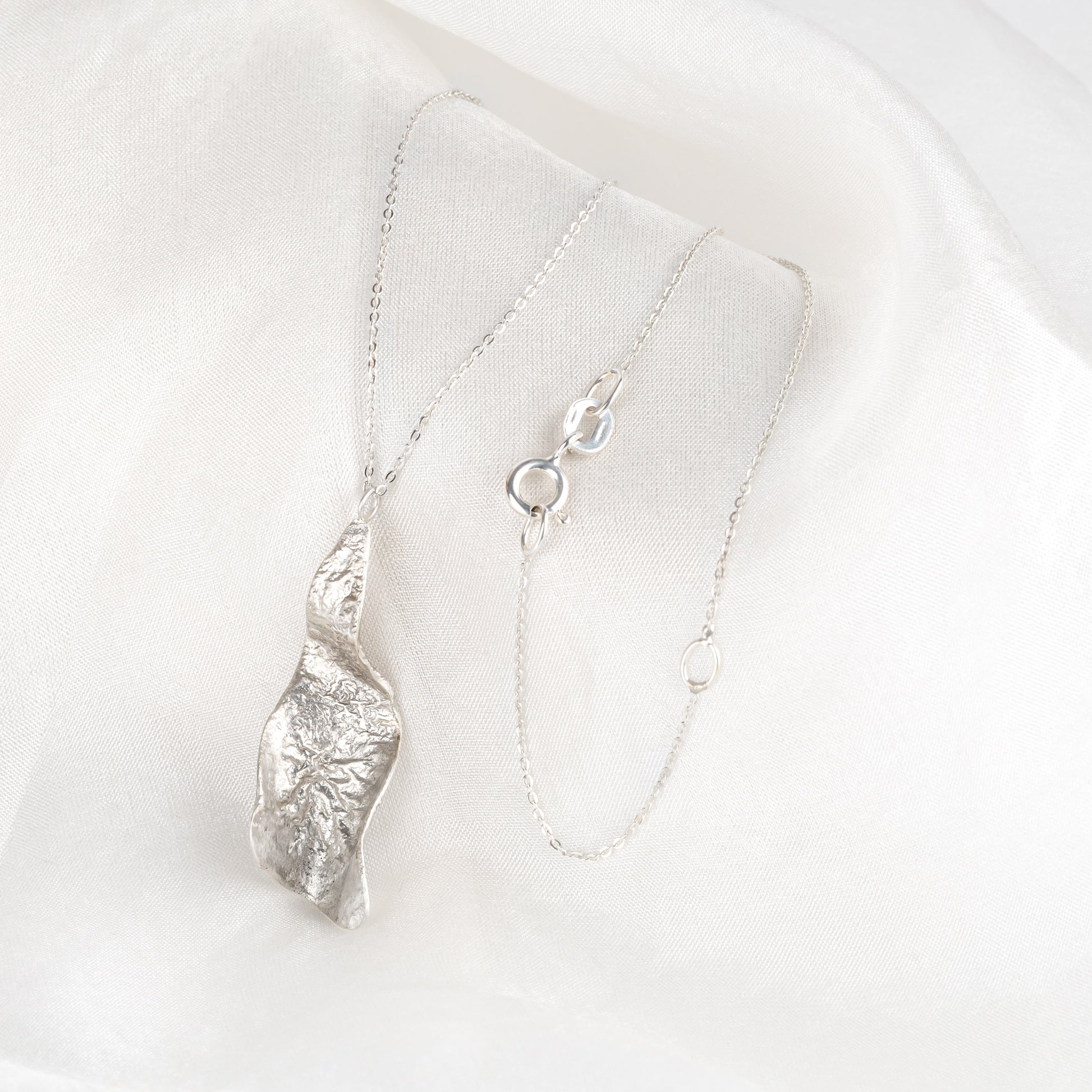 A silver textured pendant with a curled design on a chain, crafted from sterling silver draped on some fabric.