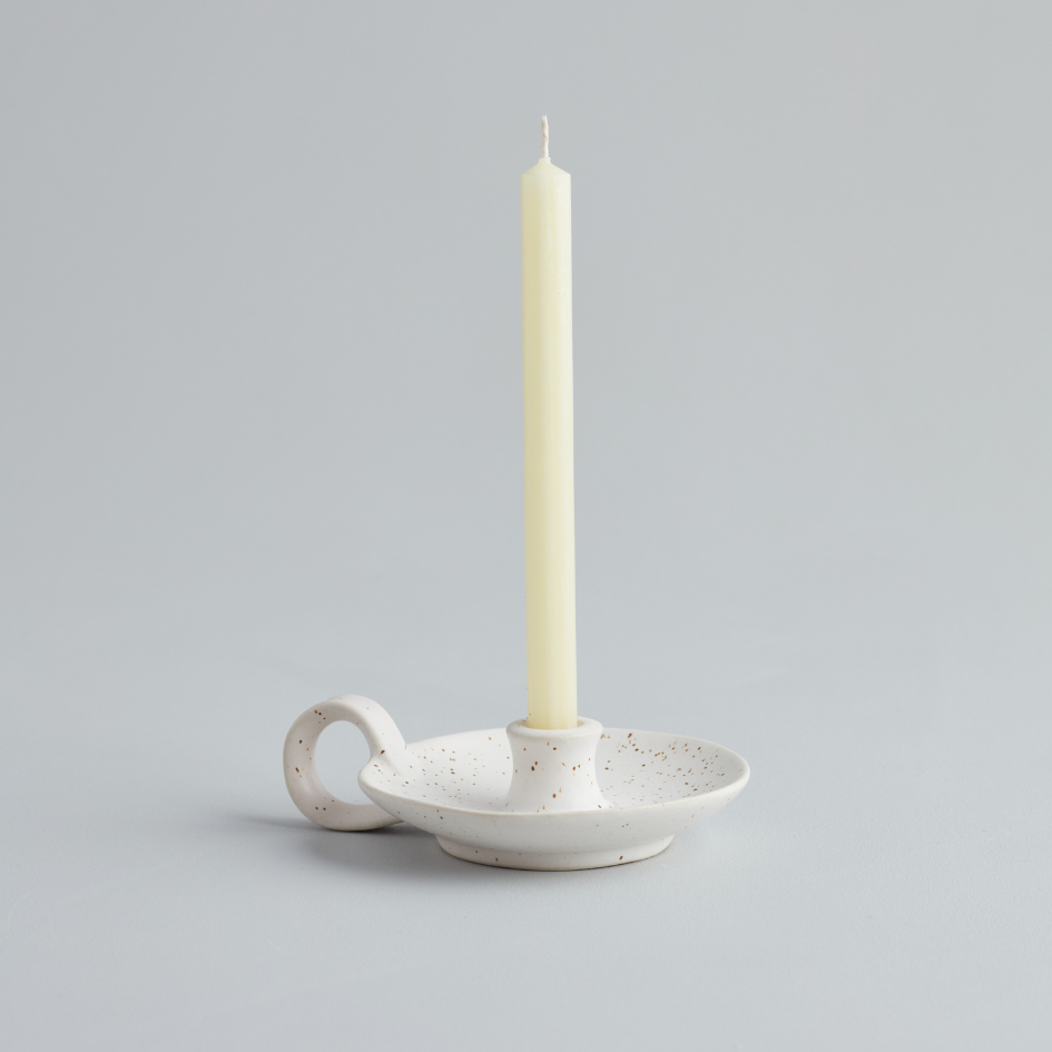 A white speckled ceramic mini candle holder with a handle, holding a plain ivory candle displayed against a plain background.