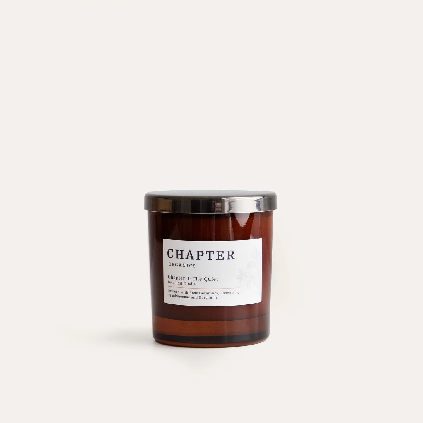 A brown glass candle jar with a silver lid, labelled "CHAPTER" and "Chapter & The Quiet". The label features a simple design with text detailing the scent, which includes notes of bergamot, rosemary, and patchouli. The background is plain and light.