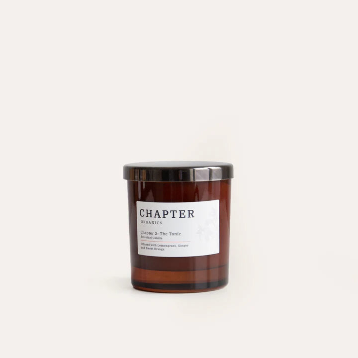 A brown glass candle jar with a black lid, labelled "Chapter Organics" and "Chapter 2: The Tonic". The label features a minimalist design with text in a simple font, set against a light, neutral background.