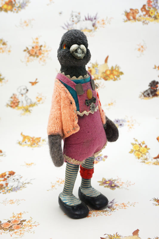 A plush pigeon dressed in a pink dress, striped leggings, and a peach cardigan with colourful buttons stands against a whimsical background featuring small animal illustrations and floral patterns.