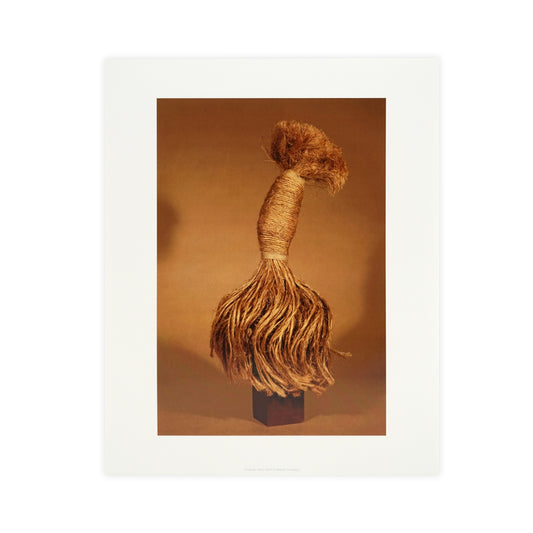 A textured, natural fibre sculpture resembling a whimsical figure, with a rounded top and flowing strands. It stands on a small wooden base against a warm, neutral background, highlighting its organic materials and artistic form.